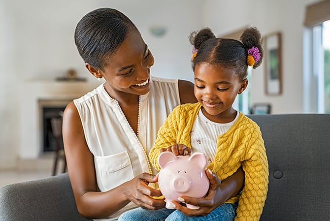 Mercado Pago: menina sentada no colo da mãe, que está segurando um cofrinho onde a criança está depositando uma moeda para começar a entender sobre educação financeira.