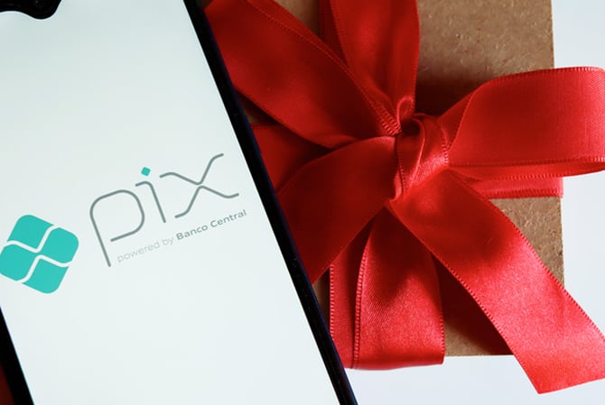 Mercado Pago: celular, com tela aberta no logo do Pix, posicionado em cima de uma caixa de presente, com laço vermelho grande, sugerindo que aquele item foi comprado graças a existência do Pix Parcelado.