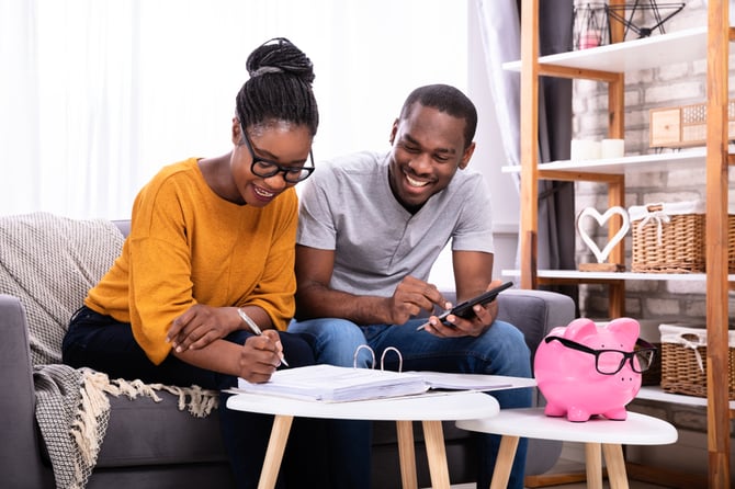 Casal sorrindo, sentado em um sofá com uma calculadora na mão e vários papéis na frente em cima de uma mesinha