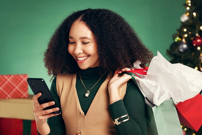 Mercado Pago: mulher em uma sala com decoração de Natal, ela está segurando sacolas e mexendo no celular para fazer mais compras de Natal.