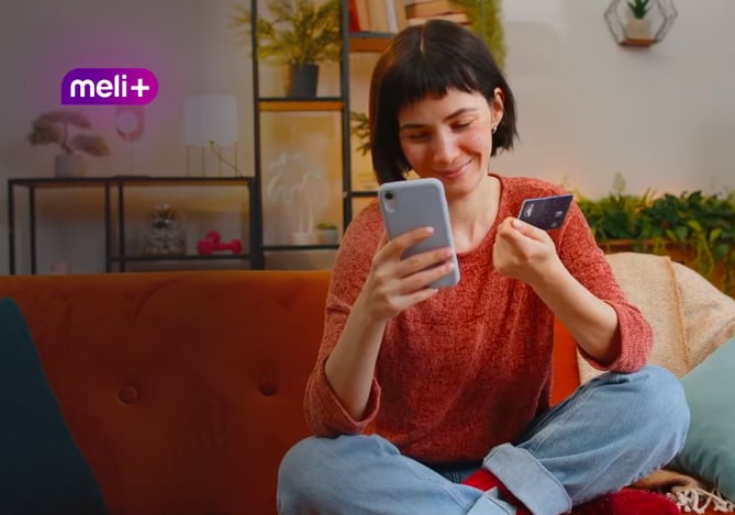 Mercado Pago: mulher sorrindo e sentada no sofá com o celular em uma mão e o cartão em outra, com o símbolo da assinatura Meli+ no canto superior esquerdo para representar a satisfação com o programa.