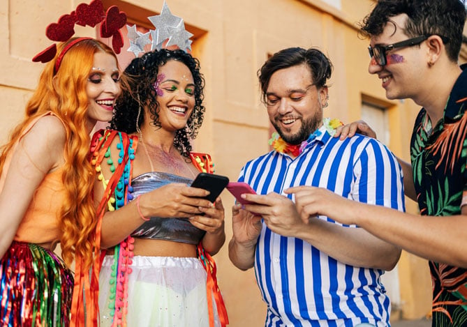 Quatro pessoas, sendo duas mulheres e dois homens, curtindo o Carnaval seguro enquanto estão fantasiadas com brilhos e serpentinas, analisando as informações do Mercado Pago no smartphone.