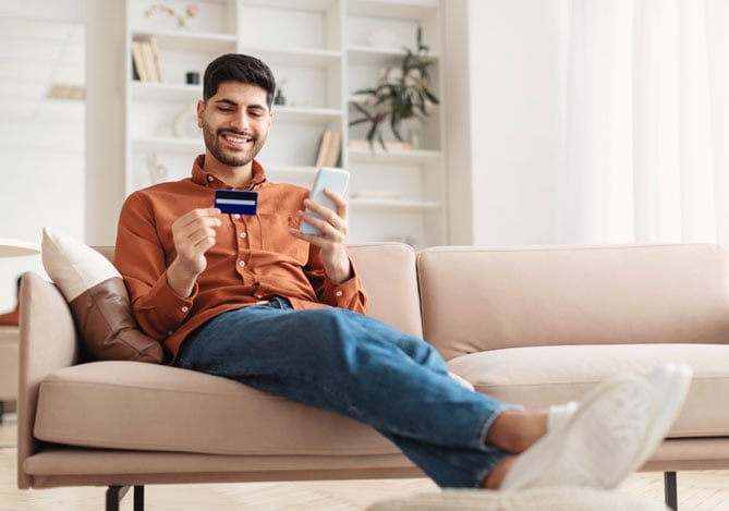 Mercado Pago: Homem sorridente sentado no sofá usando um cartão com cashback para realizar uma compra online pelo celular em um ambiente moderno e aconchegante.