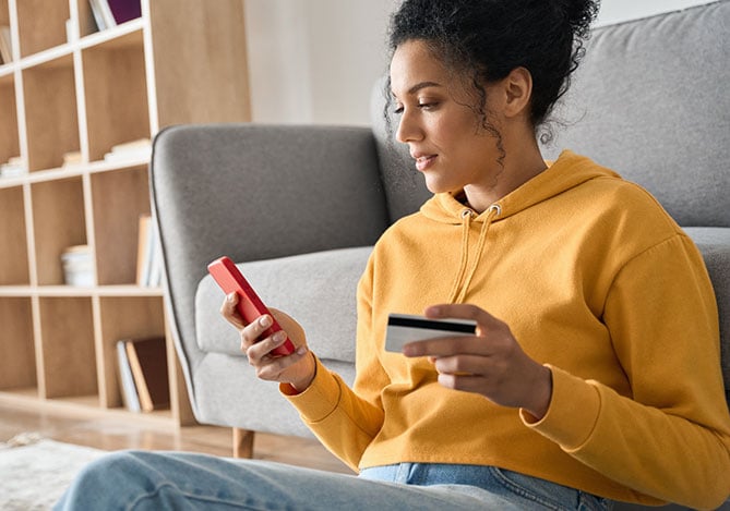 Mulher sentada no chão da sala, vestindo um moletom amarelo e calça jeans, enquanto segura o celular em uma mão e na outra o cartão de crédito para verificar sua pontuação de crédito no mercado