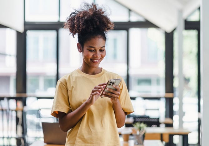Mulher com uma camiseta amarela sorrindo enquanto mexe no smartphone para acessar sua carteira digital do Mercado Pago