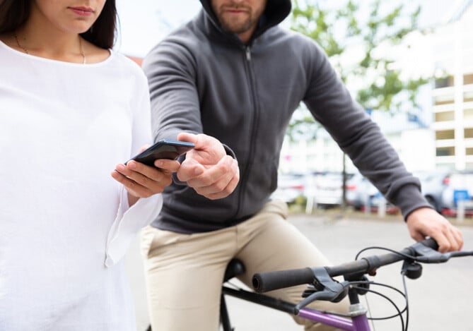 Mercado Pago: imagem de um homem em uma bicicleta com a mão esticada para pegar o celular que está nas mãos de uma mulher que caminha pela rua enquanto olha a tela para ilustrar uma situação em que o celular é roubado na rua por conta de distração.