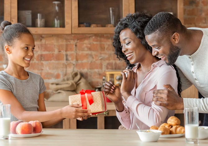 Mercado Pago: imagem de uma família celebrando o Dia das Mães. Na imagem, é possível ver a filha entregando um presente à mãe, que sorri, enquanto é abraçada pelo marido. O presente foi comprado após a garota e o pai terem pesquisado juntos formas de como economizar sem deixar de lado as boas ideias e qualidade dos presentes para as mães. na imagem, também  é possível ver que a família está na cozinha da casa e que sobre a mesa há frutas, pães e copos de leite.