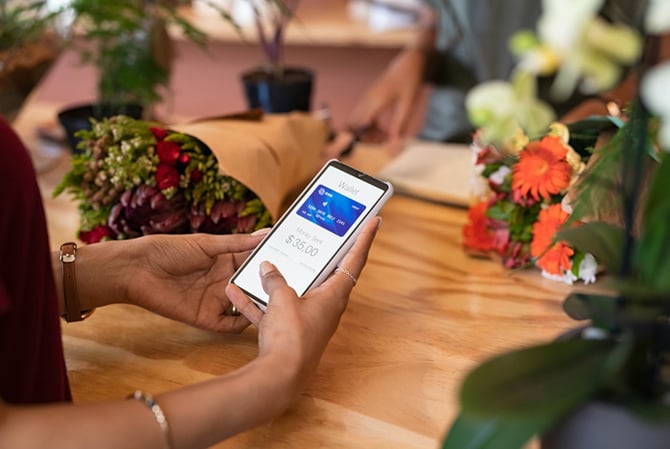 Mercado Pago, mulher sentada à mesa de restaurante com várias flores, com o celular na mão visto de cima, onde está com a carteira digital aberta representando como usar cartão virtual em loja física. 