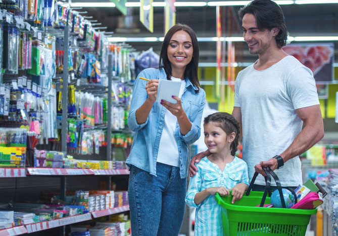 Mercado Pago: Imagem de uma família em uma papelaria, com uma lista de compras em mãos, se preparando para comprar material escolar para a filha. A mãe está de pé segurando a lista com um lápis, vestindo uma camisa e calça jeans, e camiseta branca. O pai está ao lado vestindo uma camiseta branca e segurando a cesta de compras verde e a filha está à frente dos dois, olhando para a prateleira e vestindo uma blusa xadrez verde e branca.