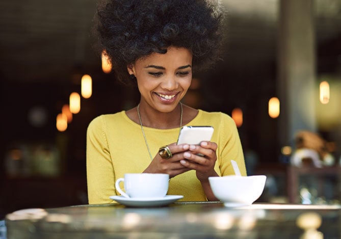 Mercado Pago: imagem de uma mulher sentada à mesa de um café enquanto acessa sua conta digital por meio do celular. Ela veste uma camiseta de manga longa amarela e sorri. Sobre a mesa, é possível ver uma xícara de café branca e um açucareiro.