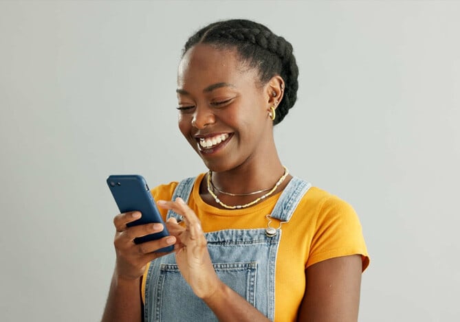 Mulher vestindo uma camiseta amarela com uma jardineira jeans segurando um celular e abrindo uma conta digital para fazer com que seu dinheiro renda mais.