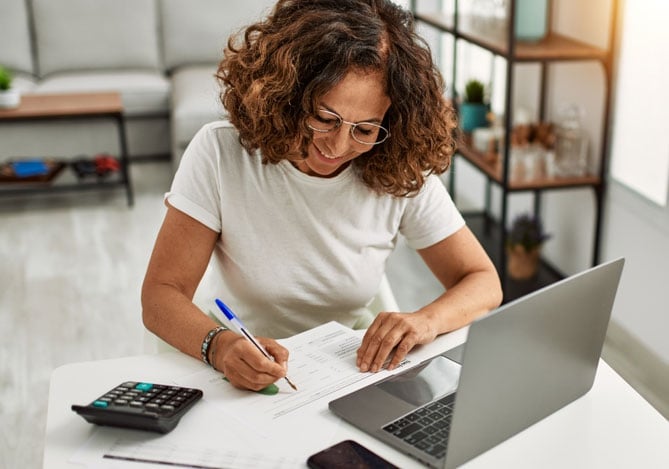Mercado Pago: Mulher analisando documentos financeiros enquanto usa um laptop, calculadora e anotações em uma mesa, simbolizando planejamento para o Dia das Mães com organização e controle de orçamento.