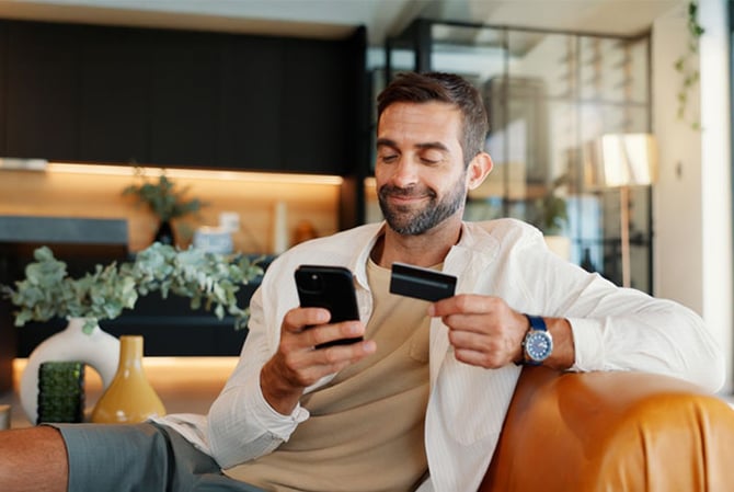 Mercado Pago: homem sentado no sofá mexendo no celular com um cartão na mão para avaliar a melhor opção de crédito para o fim do ano.