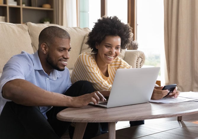 Mercado Pago: imagem de um casal sentado em frente a um notebook apoiado sobre uma mesa de centro na sala de sua casa para avaliar a possibilidade de solicitarem crédito pessoal junto a instituições financeiras. Os dois estão sorrindo, indicando que buscam crédito pessoal para a realização de algum projeto em conjunto. Enquanto o homem digita no notebook, a mulher segura um celular.