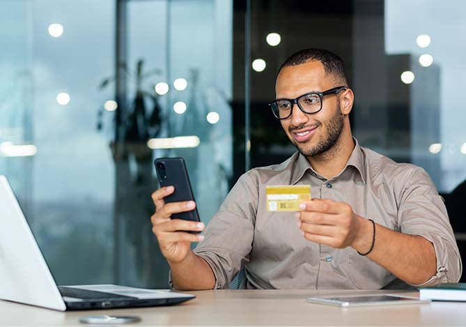 Mercado Pago: homem de óculos e camisa social mexendo no smartphone com uma mão e na outra segurando seu cartão de crédito para analisar o quanto está pagando de crédito rotativo