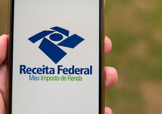 Mercado Pago: imagem de uma mão segurando um celular no qual é possível ver o logo do aplicativo da Receita Federal que os contribuintes podem acessar para fazer uma declaração pré-preenchida do Imposto de Renda 2025. Na tela do celular, é possível ler, no logo do aplicativo “Receita Federal” e, logo abaixo, o nome do app: “Meu Imposto de Renda”.