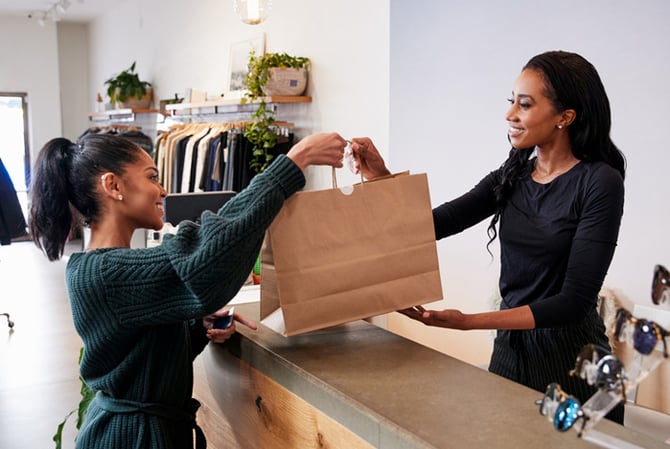 Mercado Pago: duas mulheres, uma cliente e uma vendedora, sorrindo uma para a outra, no balcão de uma loja física de roupas e acessórios, enquanto a vendedora entrega uma sacola à compradora no Dia do Cliente. 