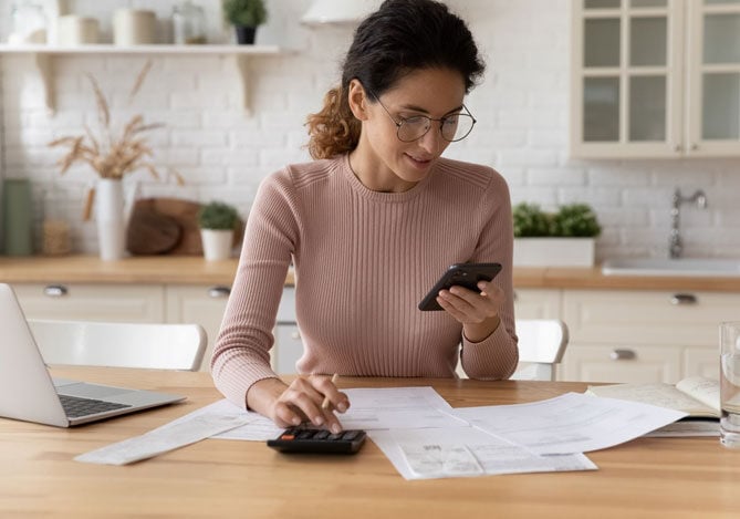 Mercado Pago: imagem de uma mulher sentada à mesa da cozinha de sua casa enquanto avalia suas opções para solicitar um empréstimo urgente. Com uma das mãos ela digita em uma calculadora, enquanto olha para a tela de um celular, que segura com a outra mão. Sobre a mesa, há um notebook aberto, algumas contas e um copo com água.
