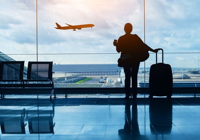 Mercado Pago: imagem da silhueta de uma pessoa na sala de embarque de um aeroporto olhando pelo vidro enquanto um avião decola. A pessoa segura um copo de bebida em uma das mãos enquanto apoia a outra mão sobre a alça do carrinho da outra mala, simulando que essa pessoa aproveitou alguma oportunidade e adquiriu passagens aéreas para fazer viagens de lazer. Na imagem também é possível ver duas das cadeiras do aeroporto.