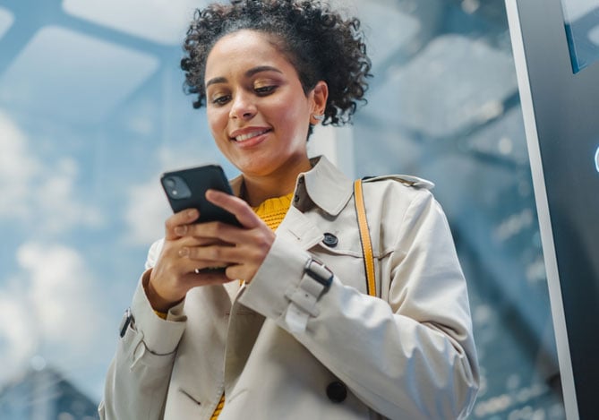 Mercado Pago: mulher sorrindo vestindo um trench coat cinza com uma blusa amarela por baixo segurando um smartphone com as duas mãos enquanto analisa as opções de financiamento