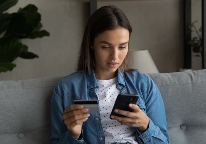 Mercado Pago: mulher sentada em um sofá com o celular em uma mão e um cartão de crédito em outra. Ela olha para a tela do dispositivo móvel de forma séria, pois desconfia que seja alvo de golpes financeiros na internet. 