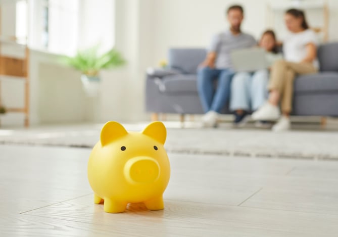Uma família formada por um homem, uma criança segurando um laptop e uma mulher, sentada em um sofá cinza no fundo da imagem e um porquinho amarelo no foco para sugerir o lançamento da nova forma de guardar dinheiro na Conta Mercado Pago, os Cofrinhos