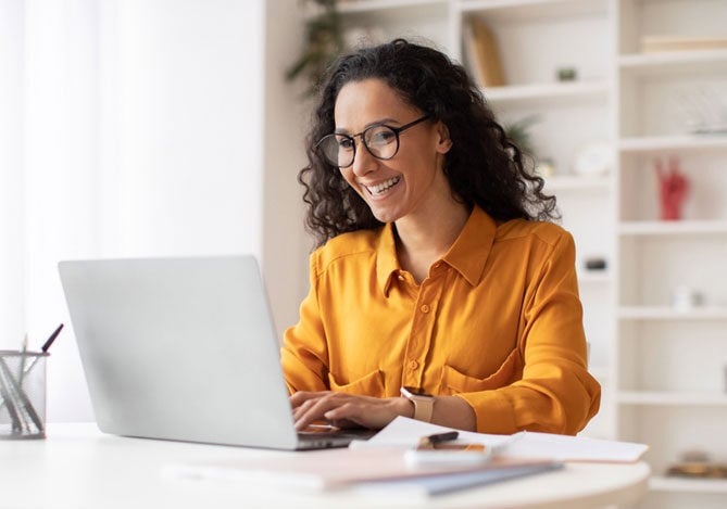 Mercado Pago: imagem de uma mulher vestindo camisa de manga longa laranja e sorrindo enquanto consulta seu holerite online usando um notebook. A mulher está usando o notebook em sua mesa de trabalho, sobre a qual também é possível ver alguns papéis e canetas.