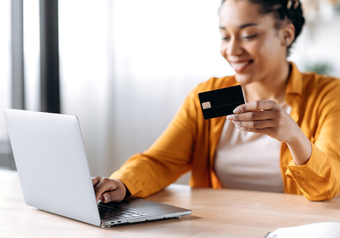 Mercado Pago: imagem de uma mulher usando um notebook e segurando um um cartão de crédito nas mãos enquanto pesquisa sobre formas de reduzir o IOF sobre suas operações de crédito.