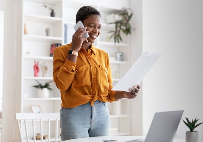 Mulher vestindo uma camisa amarela com calça jeans, falando no celular enquanto avalia sua independência financeira Mercado Pago em um papel