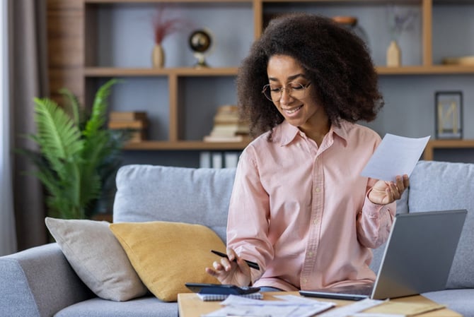 Mercado Pago: mulher sentada no sofá de sua casa, com notebook, papéis e caneta na mesinha de centro, enquanto ela se planeja para conquistar sua independência financeira.