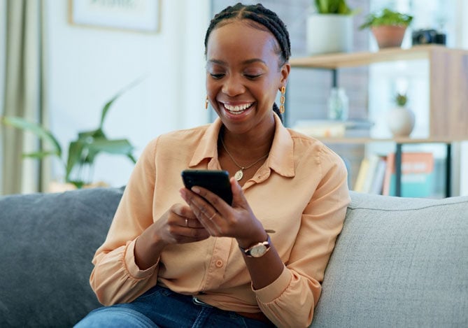 Mercado Pago: imagem de uma mulher sorrindo vestindo camisa salmão de manga longa sentada no sofá de sua casa enquanto utiliza o celular para verificar as melhores opções de investimento com uso de Inteligência Artificial. 