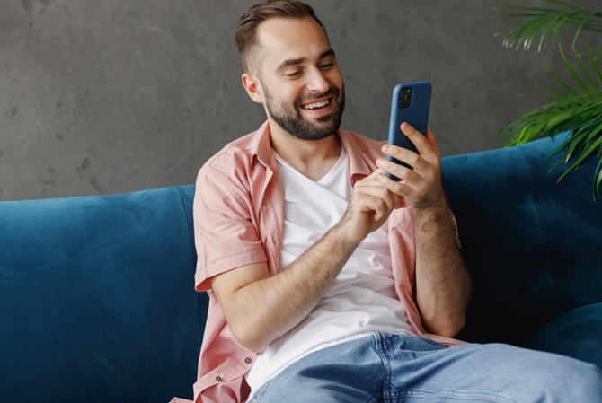 Mercado Pago: homem sentado no sofá, sorrindo e mexendo no celular para aplicar seu dinheiro em um investimento de curto prazo.