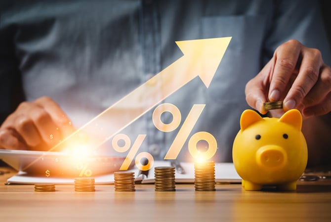 Mercado Pago: pessoa sentada atrás de uma mesa onde tem pilhas de moedas, um cofrinho e ilustrações de setas e porcentagem, representando o estudo em um investimento de renda fixa.