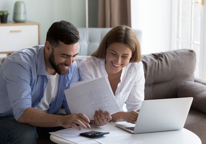 Mercado Pago: imagem de um casal sentado no sofá de sua casa, debruçados sobre uma mesa de centro onde há uma calculadora e um notebook aberto, os quais são usados para que o casal possa analisar o que pode ser mais interessante para eles: investir ou quitar as dívidas que possuem.