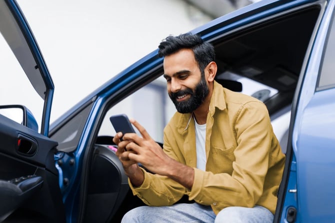 Mercado Pago: homem sentado em um carro com a porta aberta, segurando um celular nas mãos, realizando o licenciamento de veículo pela internet. 