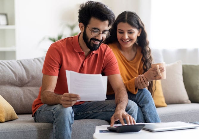 Mercado Pago: imagem de um casal sentado no sofá de casa avaliando qual pode ser a melhor opção para seus planos financeiros entre o limite de crédito e um empréstimo pessoal. O rapaz, que veste camisa vermelha, segura folhas em uma das mãos, enquanto usa uma calculadora com a outra mão. A moça, que veste camisa de manga longa amarela, sorri, enquanto olha para os papéis.