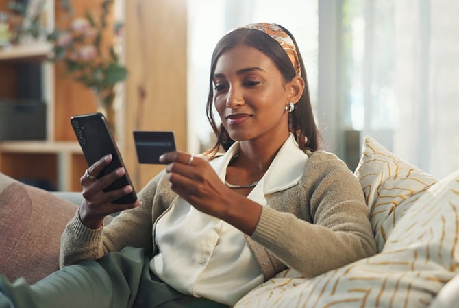Mercado Pago: mulher sentada no sofá, com o celular em uma mão e o cartão em outra, avaliando como aumentar o limite do cartão de crédito. 