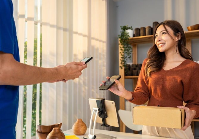 Mercado Pago: duas pessoas em um escritório, sendo um homem com celular na mão para escolher a forma de pagamento que será realizada para a mulher, que também está com seu celular em uma mão e segurando uma caixa na outra mão.