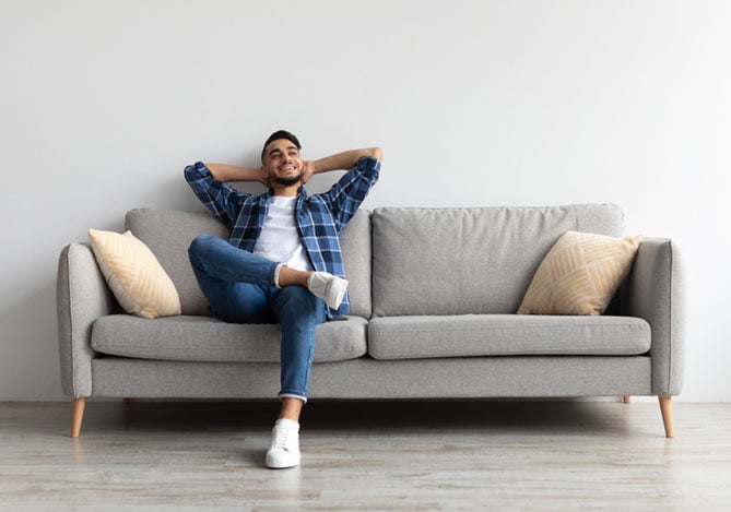 Homem sentado de pernas cruzadas no sofá de sua casa após decidir morar sozinho com auxílio das dicas de planejamento financeiro do Mercado Pago
