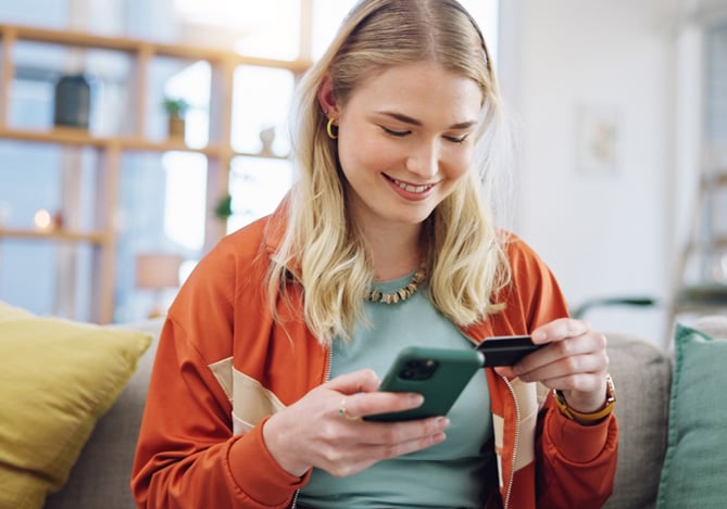 Mercado Pago: Mulher sorridente sentada no sofá usando o celular e segurando um cartão, representando a praticidade de pagar boleto com cartão de crédito pelo app.