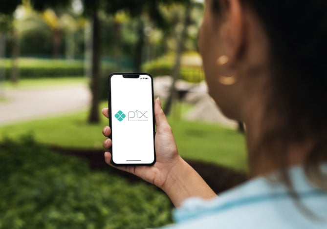 Mercado Pago: mãos de uma mulher que veste uma camisa azul, argola dourada e cabelo preso, segurando o smartphone com a tela aberta no logo do Pix, para falar das declarações de Pix no Imposto de Renda