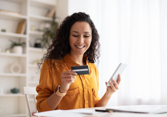 Mercado Pago: mulher sorrindo de cabelo enrolado vestindo camisa na cor amarela segurando um cartão de crédito na mão e na outra o celular para fazer o pix parcelado