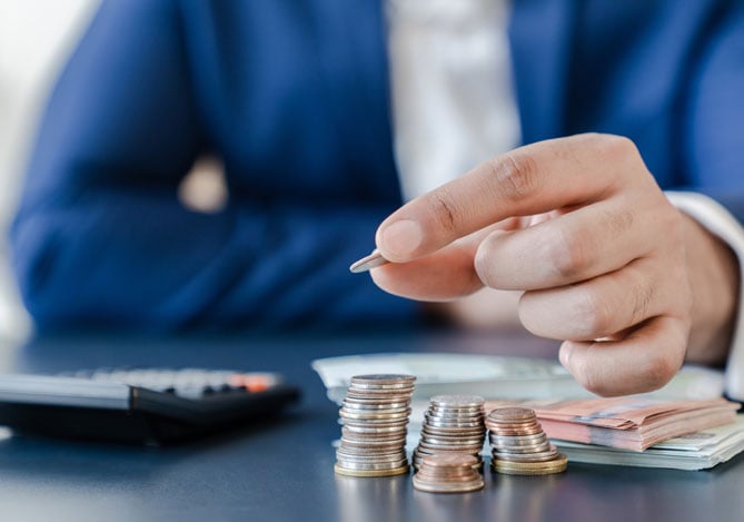 Mercado Pago: imagem da mão de uma pessoa colocando moedas em uma pilha de dinheiro sobre uma mesa onde também há cédulas e uma calculadora, ilustrando a ação de planejar uma reserva financeira para emergências ou projetos pessoais.