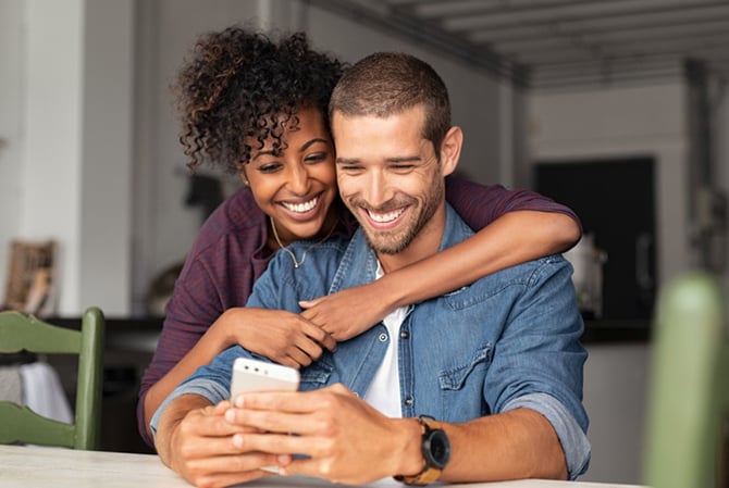 Sala de estar de uma casa e um casal. A mulher em pé, negra de cabelo pretos cacheados e preso, sorrindo e vestindo uma camisa roxa, abraçando o homem, que está sentado na cadeira, sorrindo, vestindo uma camiseta branca e uma camisa jeans por cima, além de relógio em um dos braços, segurando um celular, posicionando ele para registrar uma foto que demonstra o quanto a saúde emocional de ambos está em equilíbrio com o seu financeiro graças as soluções do Mercado Pago.