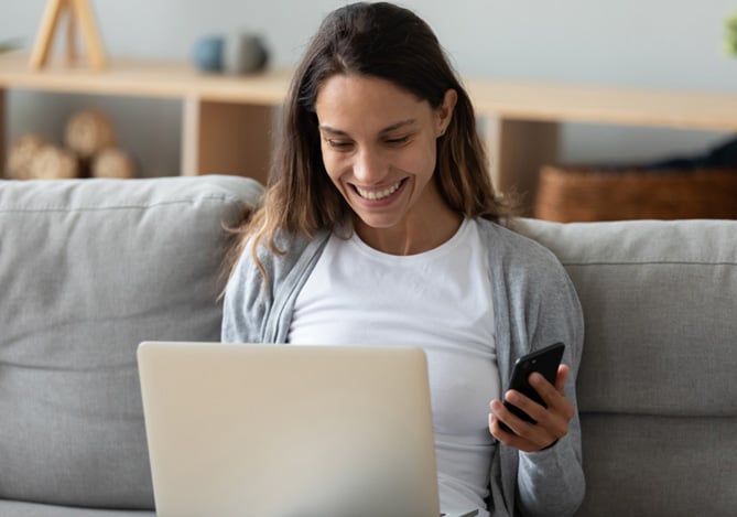 Mulher sorrindo enquanto mexe no computador e celular, identificando todas as soluções de segurança que o Mercado Pago oferece para ela.