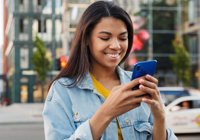 Mulher na rua, vestindo uma jaqueta jeans, enquanto mexe no celular para ver as soluções do Mercado Pago