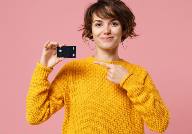 Mulher de cabelo castanhos curto, com brinco no formato de argolas grandes, vestindo suéter amarelo segurando o Cartão Mercado Pago em uma das suas mãos e a outra apontando para o cartão com o intuito de mostrar a todos as vantagens do Cartão Mercado Pago.