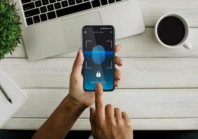 Mercado Pago: mesa de madeira com um laptop aberto, caderno, lápis, xícara com café e planta, e acima disso, tem duas mãos de uma pessoa segurando um smartphone solicitando a biometria para destravar e assim evitar o roubo de identidade online.