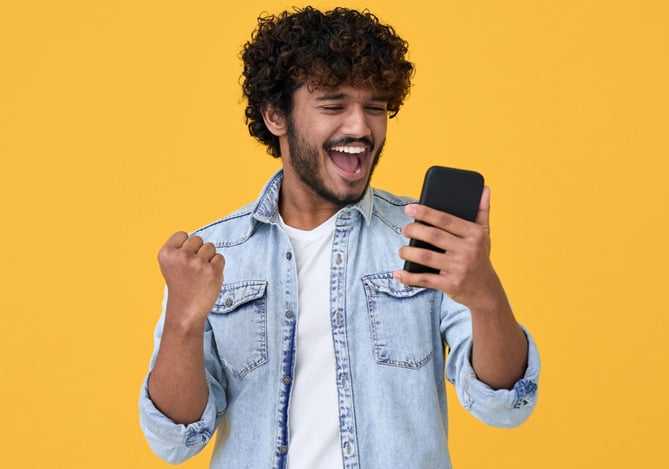 Homem preto vestindo uma camiseta branca e por cima uma jaqueta jeans celebrando enquanto olha seu celular com informações sobre rendimento Mercado Pago