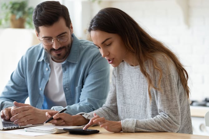 Casal com camisa azul e blusa cinza sentando em mesa de escritório e com uma calculadora declarando seu Imposto de Renda com ajuda do Mercado Pago